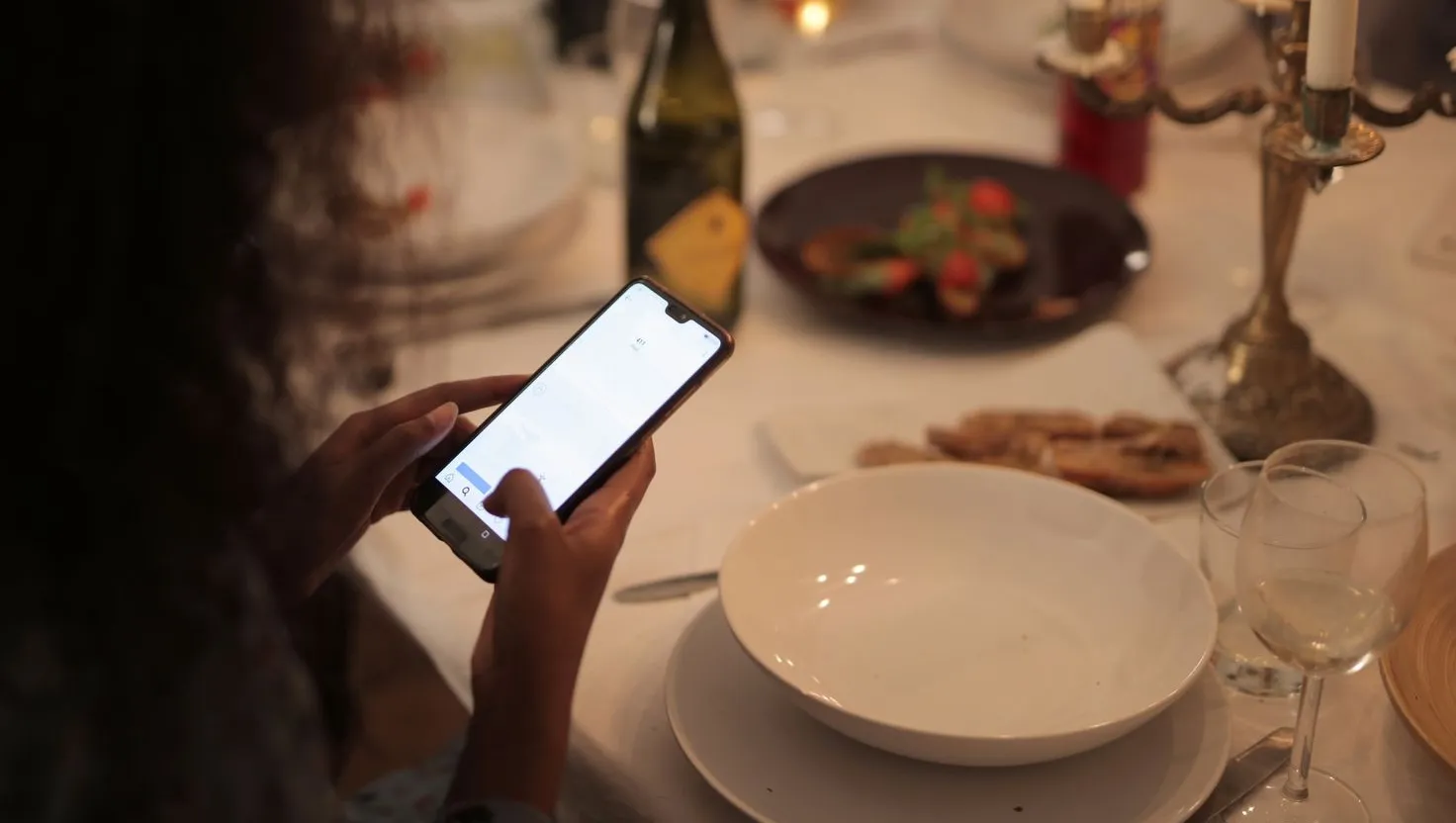 Cliente usando o celular numa mesa de restaurante posta, com pratos, taças e castiçal
