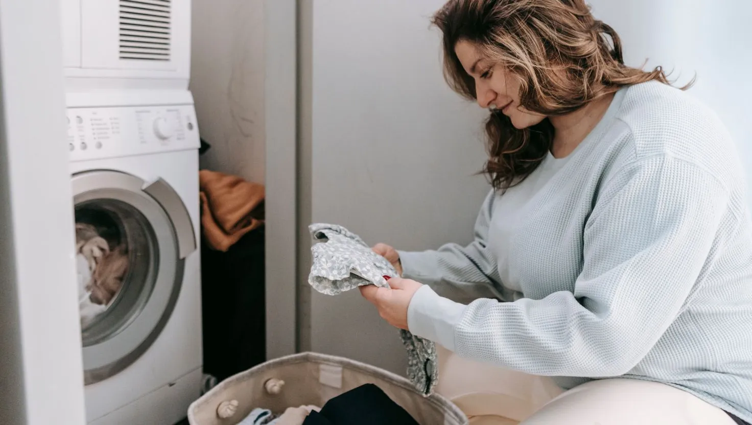 Mulher em casa separando roupas ao lado da máquina de lavar, com um cesto de roupa à frente