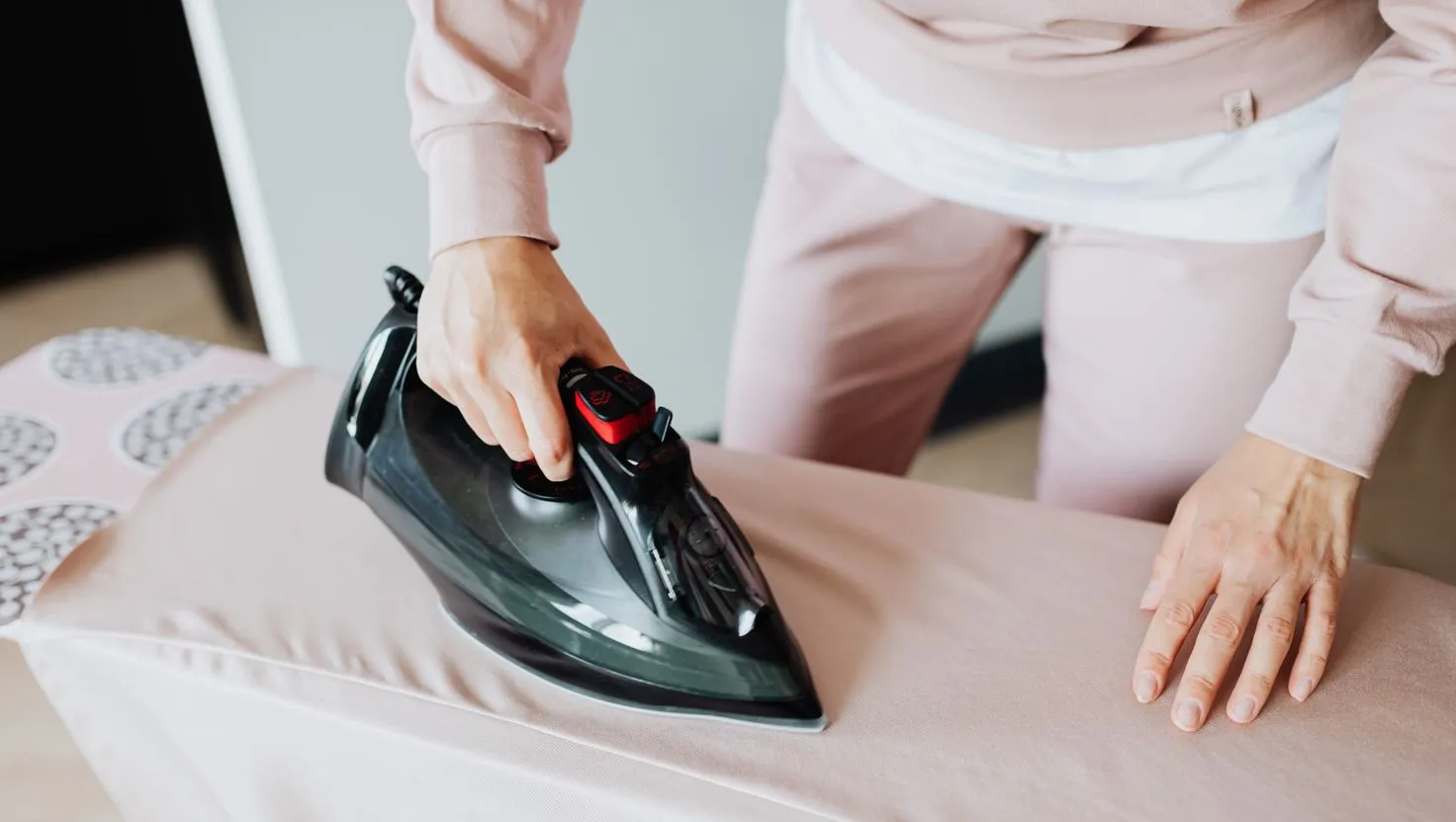 Mãos de uma pessoa passando um tecido rosa com um ferro a vapor preto sobre a tábua de passar