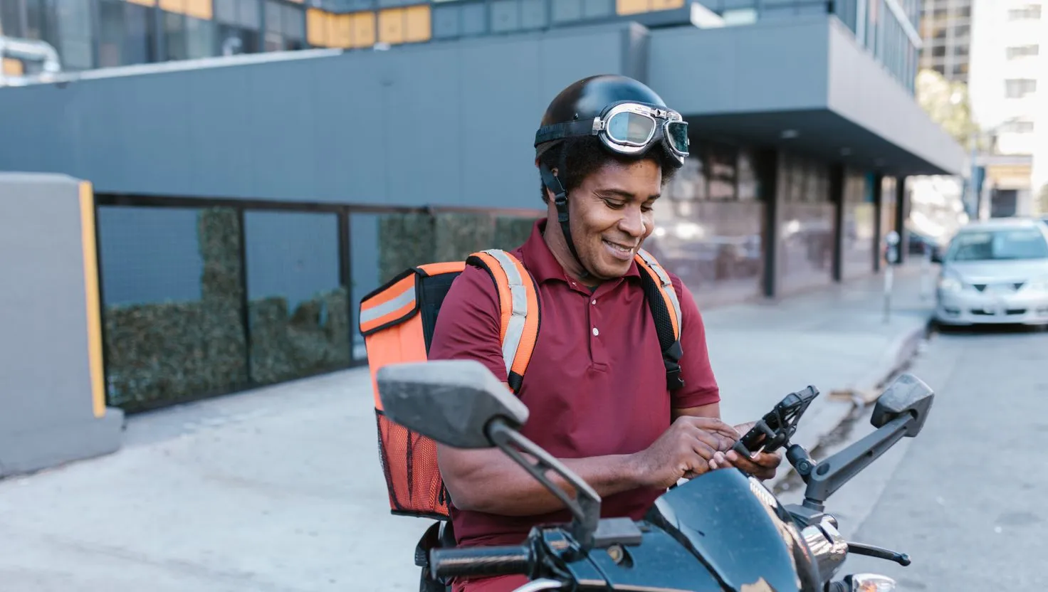 Entregador sorridente sentado na moto, com mochila de entrega laranja nas costas, conferindo o celular preso ao guidão