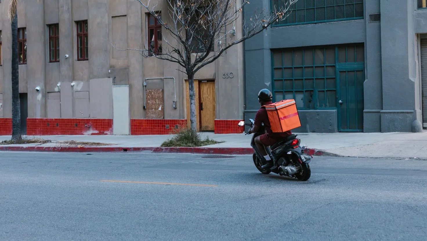 Entregador de scooter com baú térmico laranja atravessando uma esquina da cidade